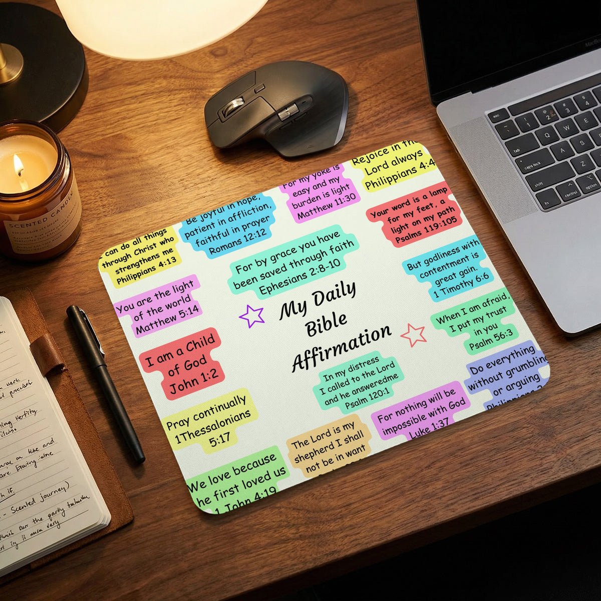 A mouse pad featuring colorful boxes with Bible verses is set on a wooden desk next to a laptop, mouse, candle, and notebook.