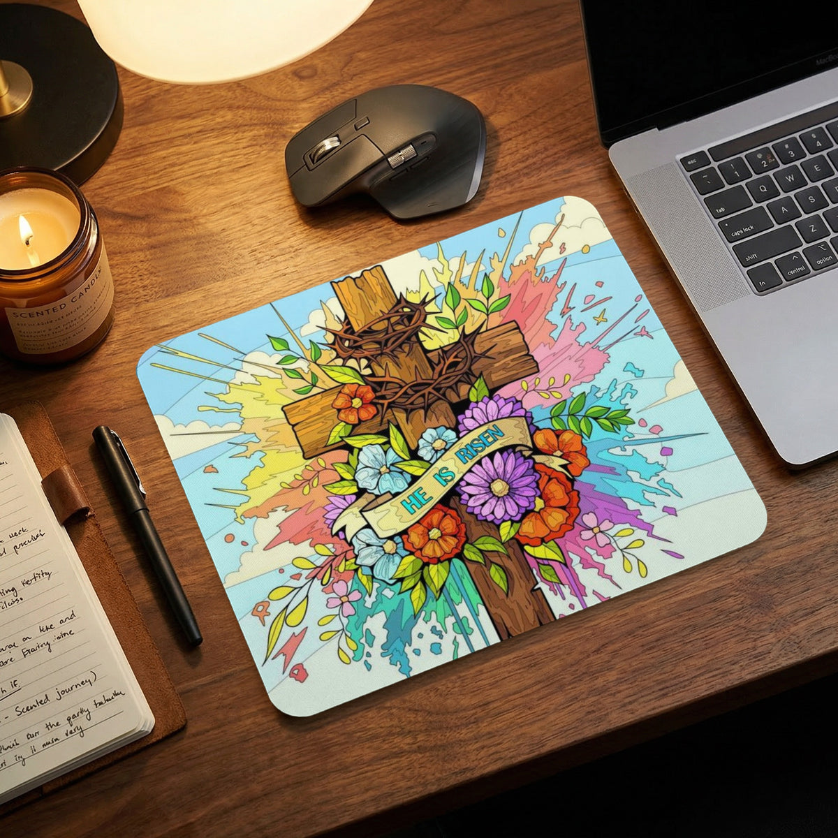 A rectangular mousepad featuring a cross, flowers, and the text He Is Risen rests on a wooden desk.