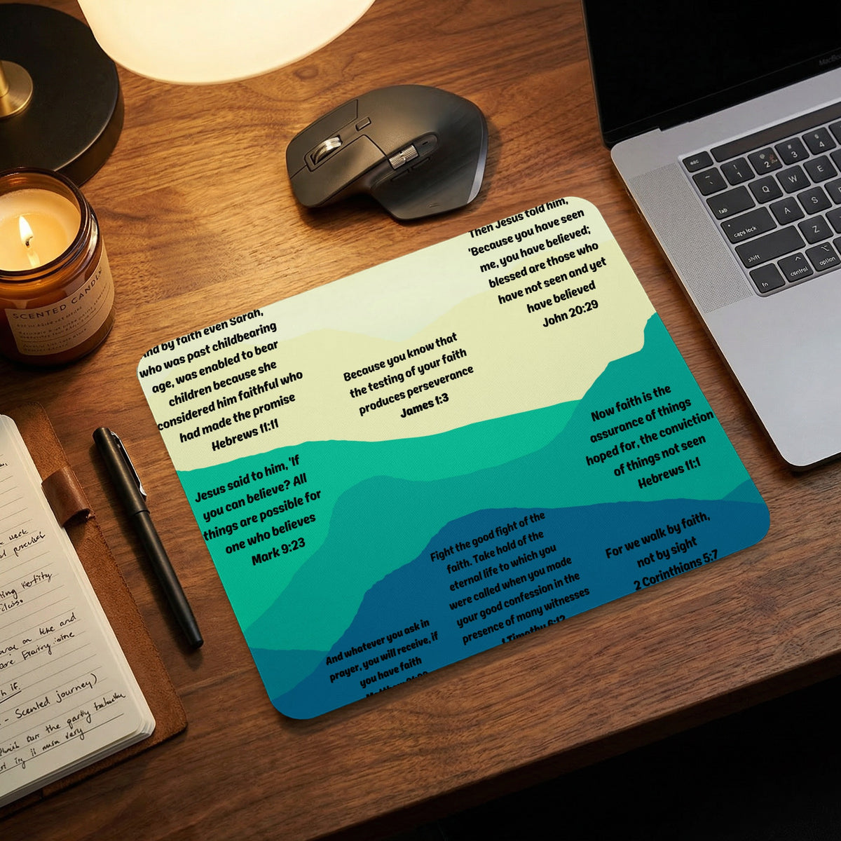 A mousepad with abstract green hills and Bible verses rests on a wooden desk next to a laptop.