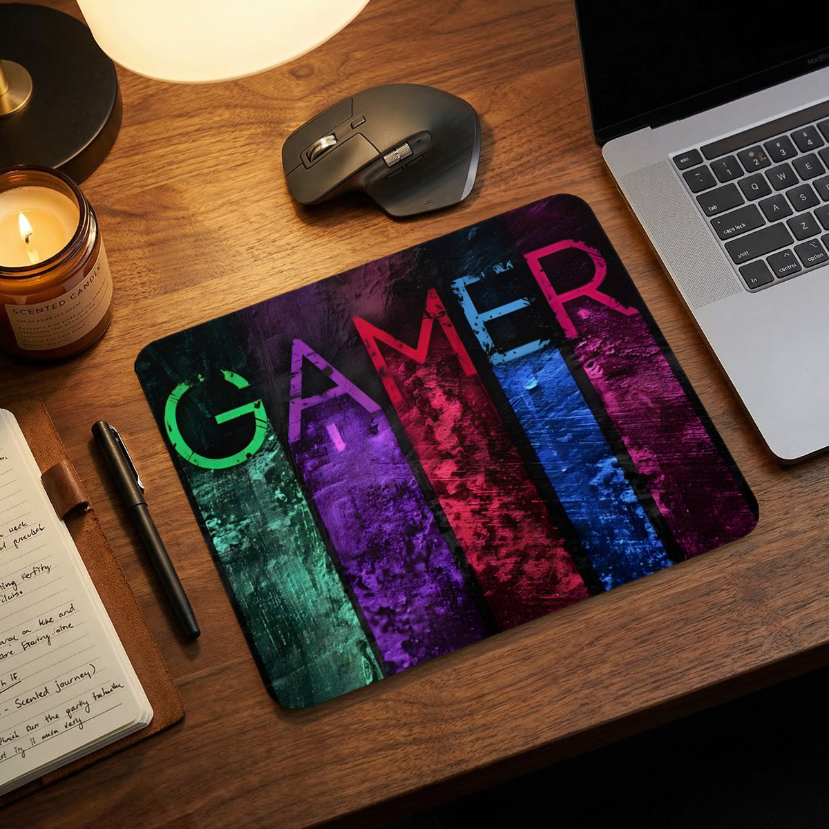 A rectangular mousepad featuring the word GAMER in vibrant, multicolored vertical stripes on a wooden desk.