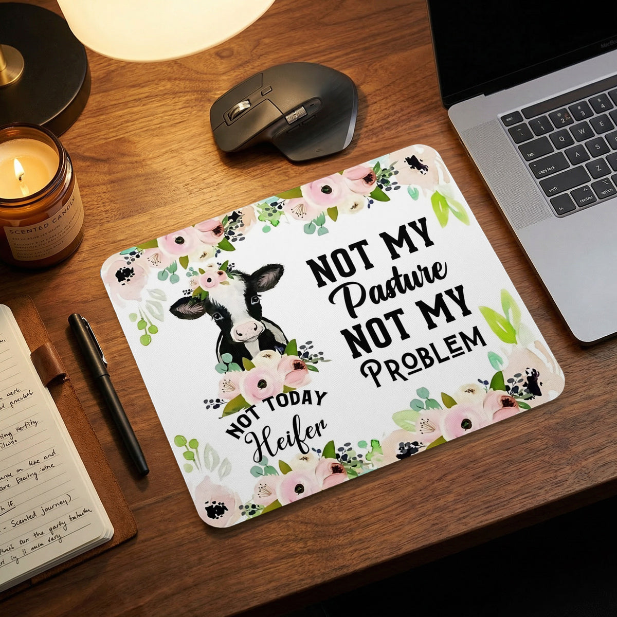 A mouse pad featuring a cow illustration, floral accents, and the text Not My Pasture Not My Problem on a wooden desk.