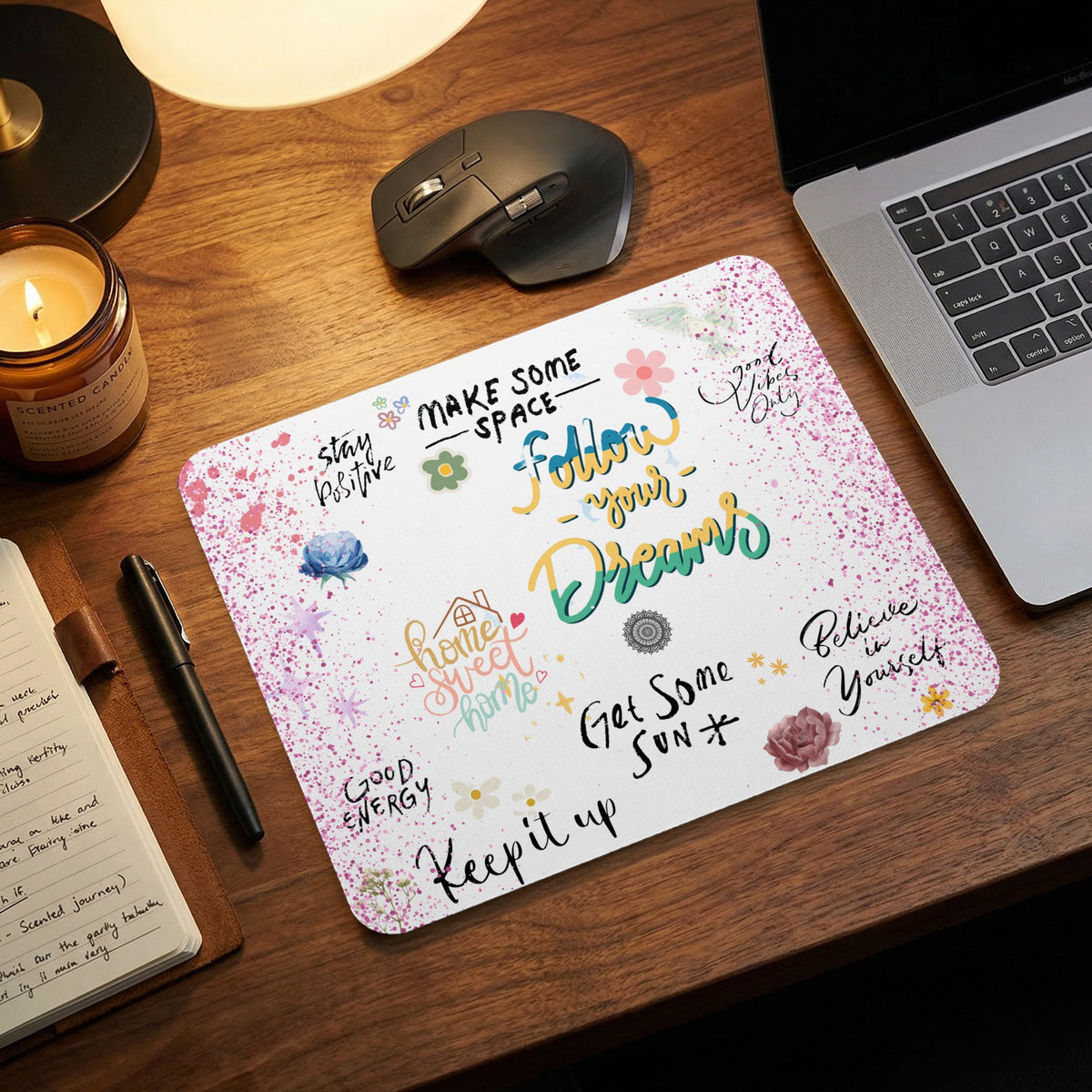 A rectangular mousepad featuring motivational quotes and pink watercolor splashes, resting on a wooden desk.