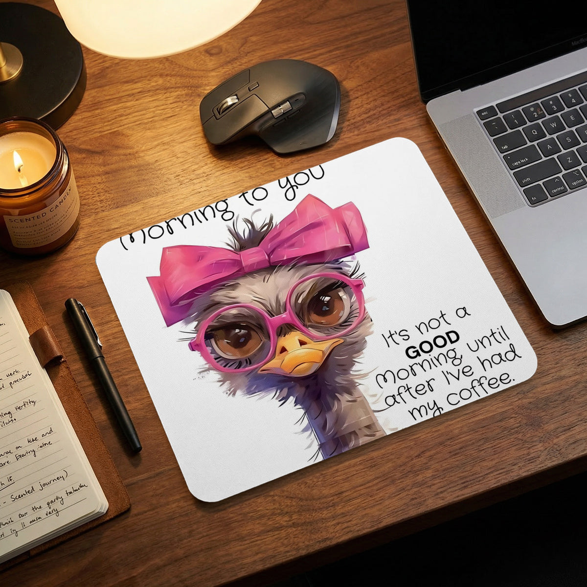 An ostrich mousepad with pink glasses and bow, featuring coffee humor text, sits on a wooden desk.