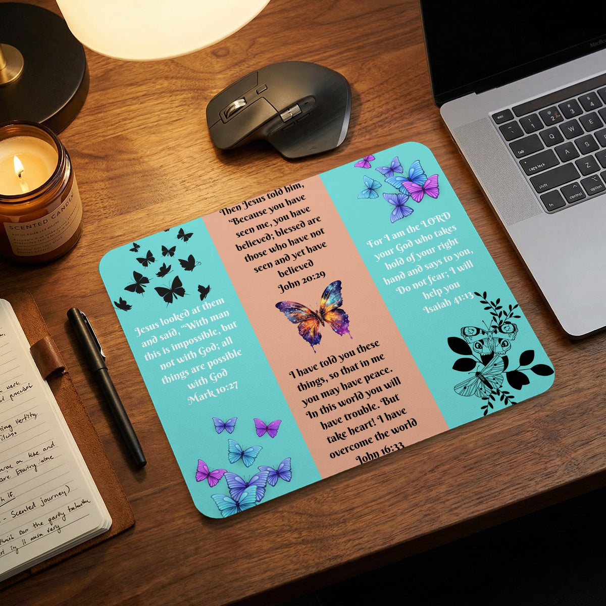A desk mat featuring colorful butterflies and Bible verses rests on a wooden desk with a laptop, mouse, and candle.