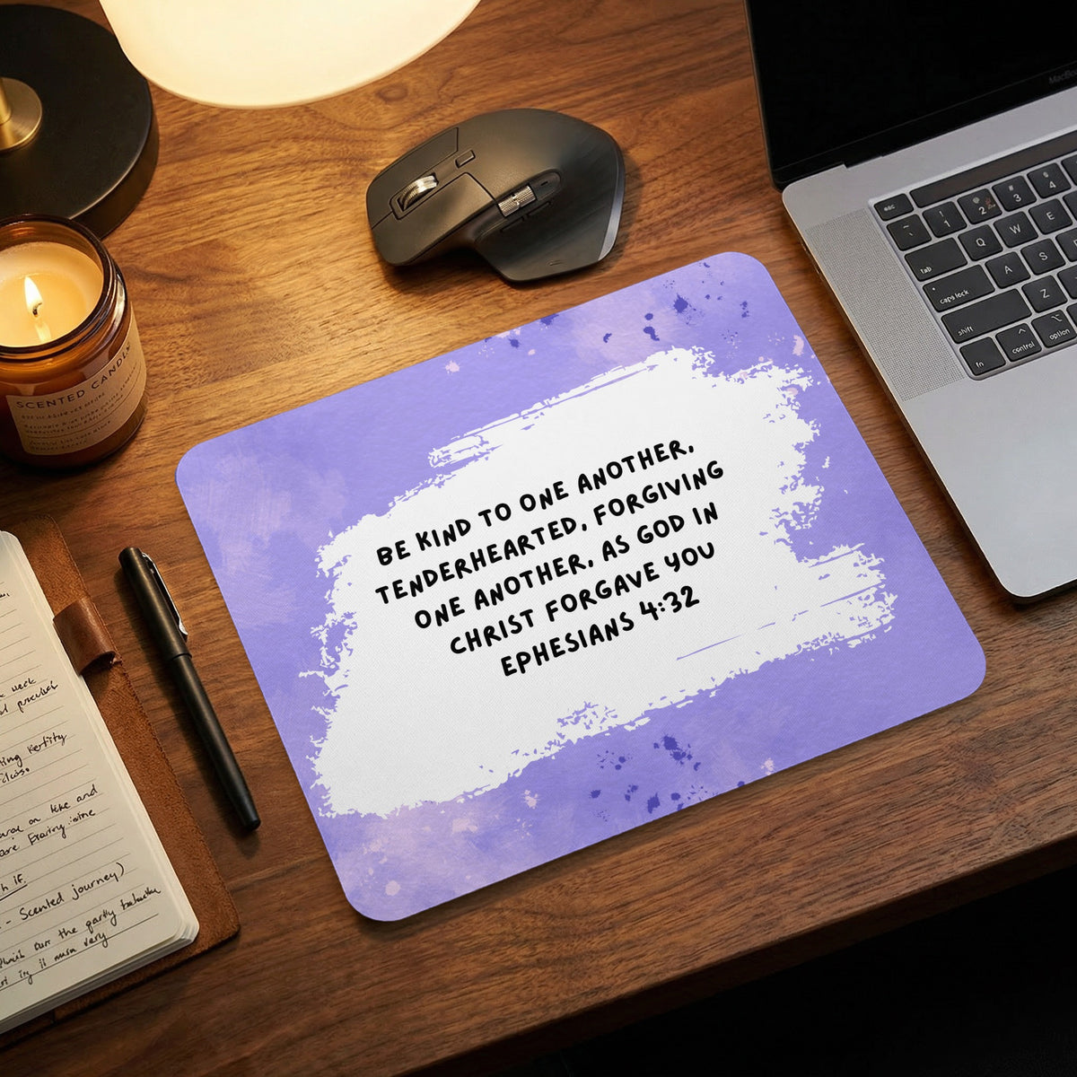 A purple mousepad featuring the Bible verse Ephesians 4:32 rests on a wooden desk with a laptop and notebook.