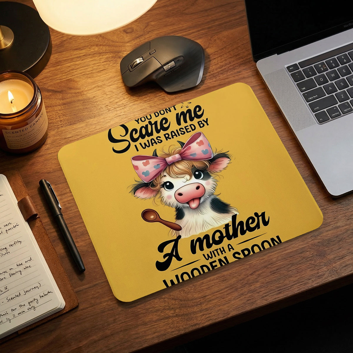 A yellow mouse pad on a wooden desk with a cow design and the text You dont scare me I was raised by a mother with a wooden spoon.