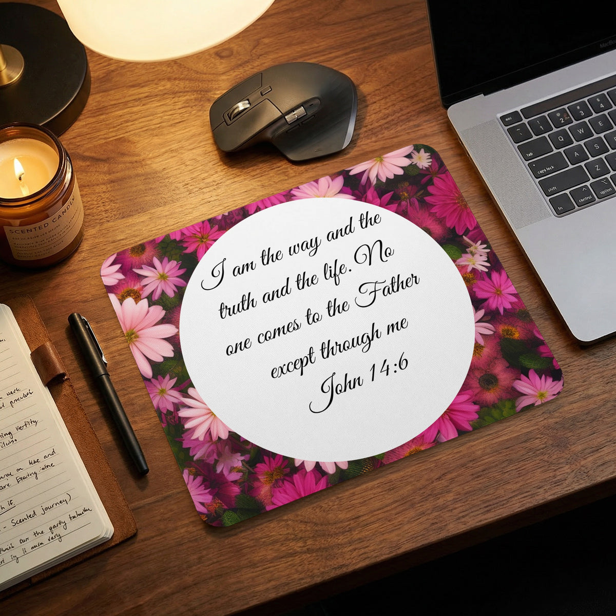 A rectangular mousepad featuring pink daisies and the bible verse John 14:6 rests on a wooden desk near a laptop, mouse, and notebook.