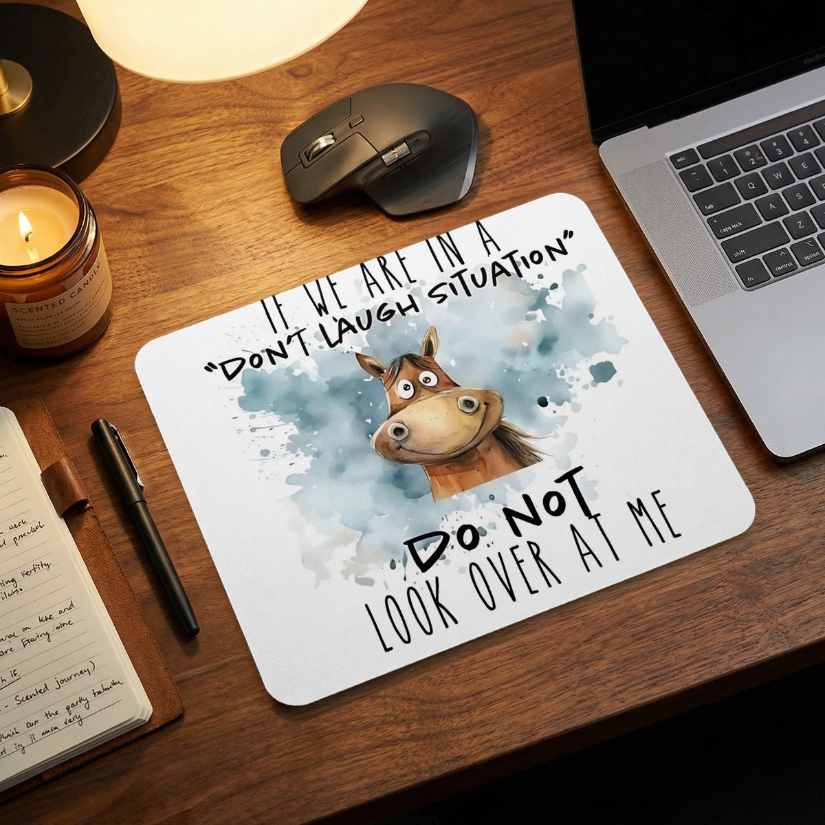 A white mousepad on a wooden desk features a whimsical cartoon horse and humorous text.