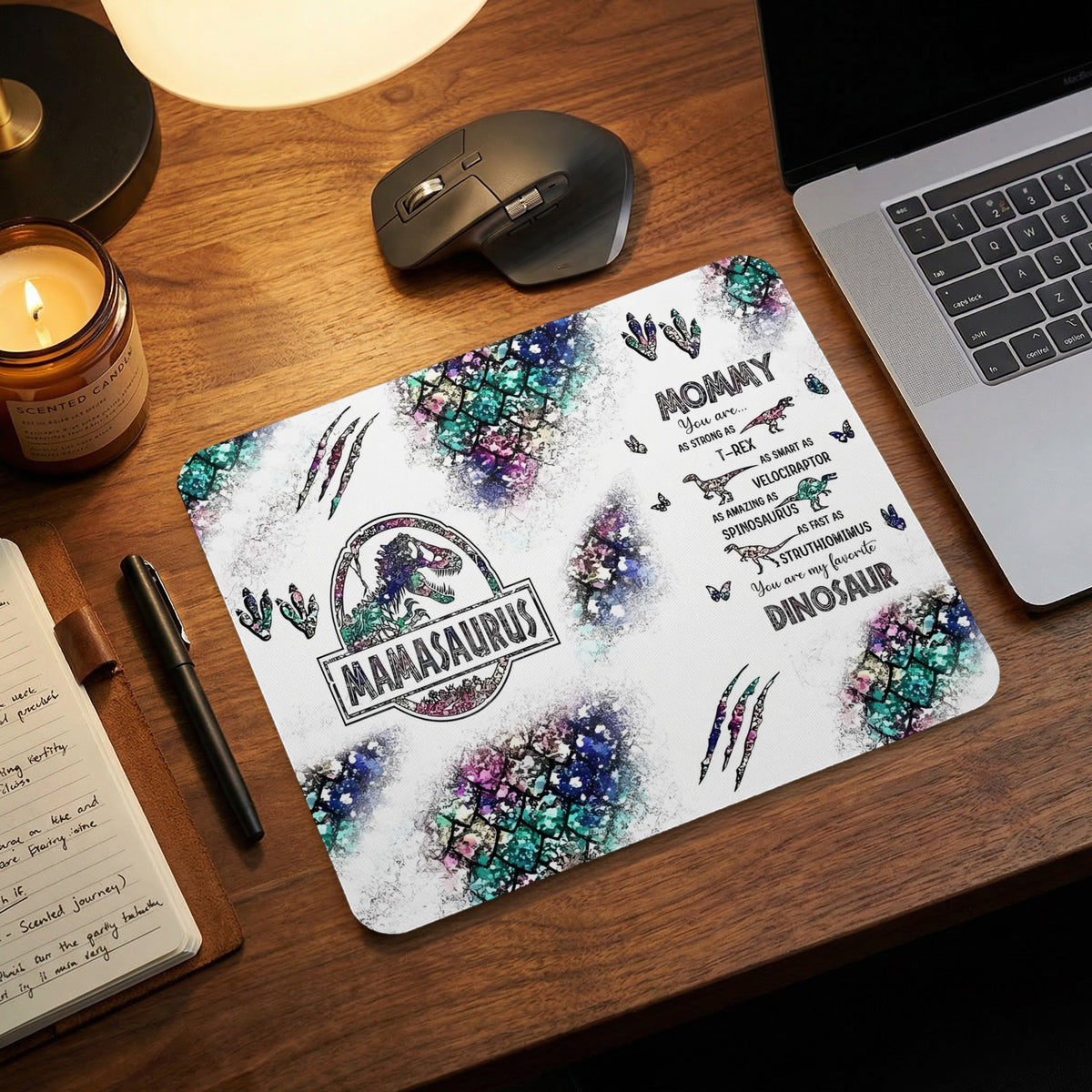 A rectangular mousepad featuring a Mamasaurus design, watercolor dinosaur scales, and text on a wooden desk.