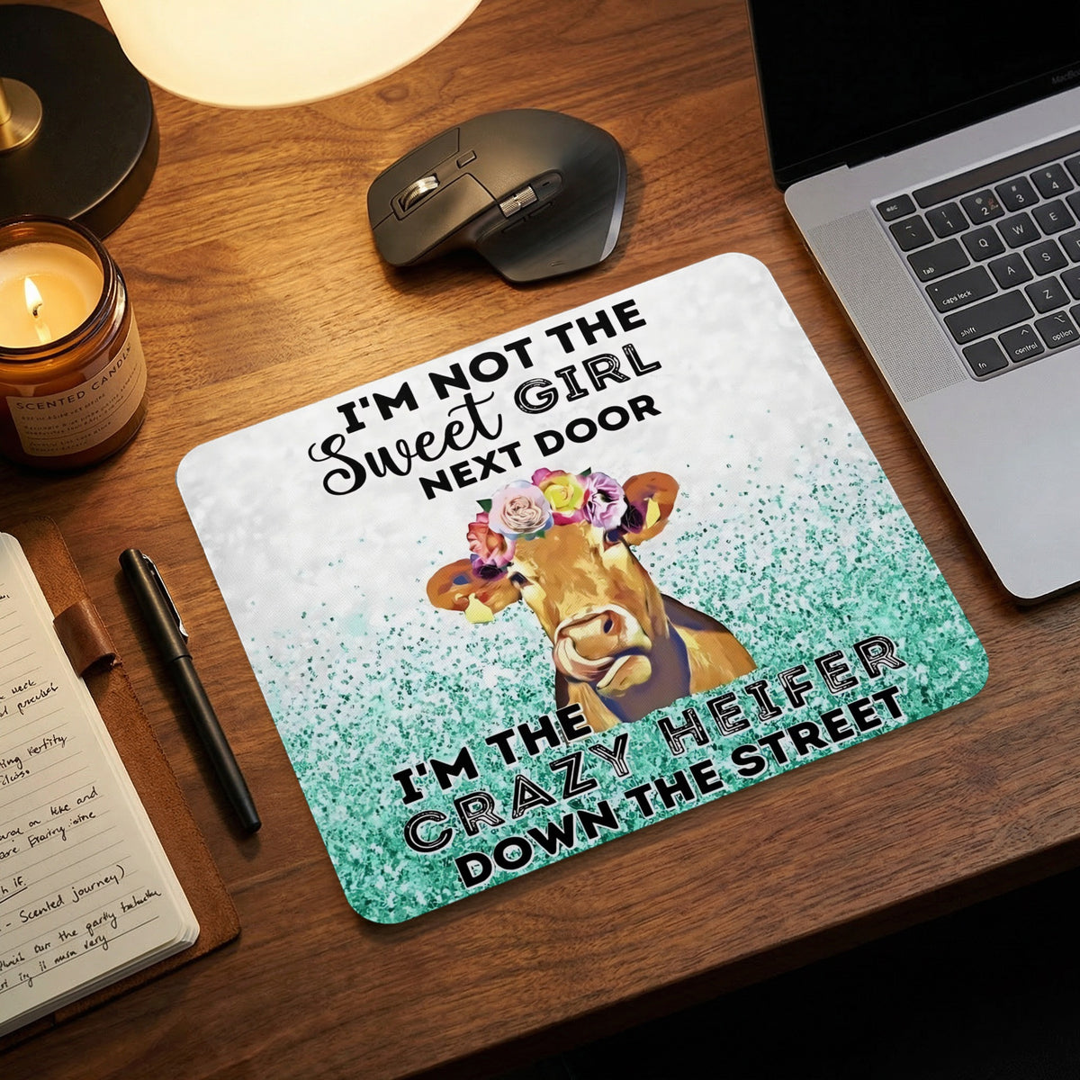 A rectangular mouse pad with a cow wearing a flower crown and text reading I AM NOT THE SWEET GIRL NEXT DOOR I AM THE CRAZY HEIFER DOWN THE STREET sits on a wooden desk.
