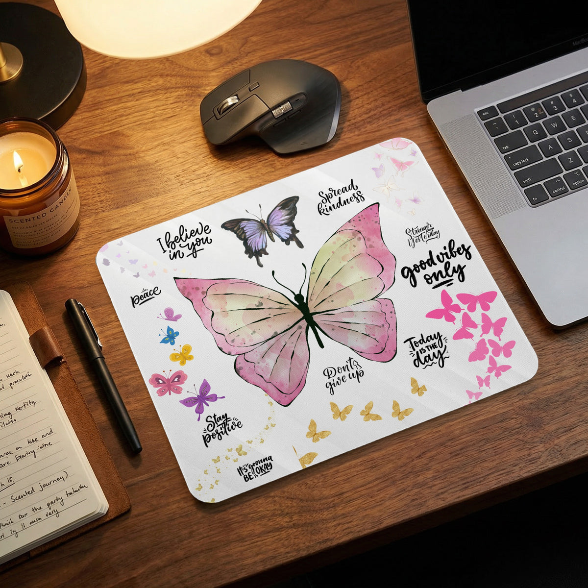 A rectangular mousepad featuring watercolor butterflies and motivational phrases sits on a wooden desk near a laptop, mouse, and candle.