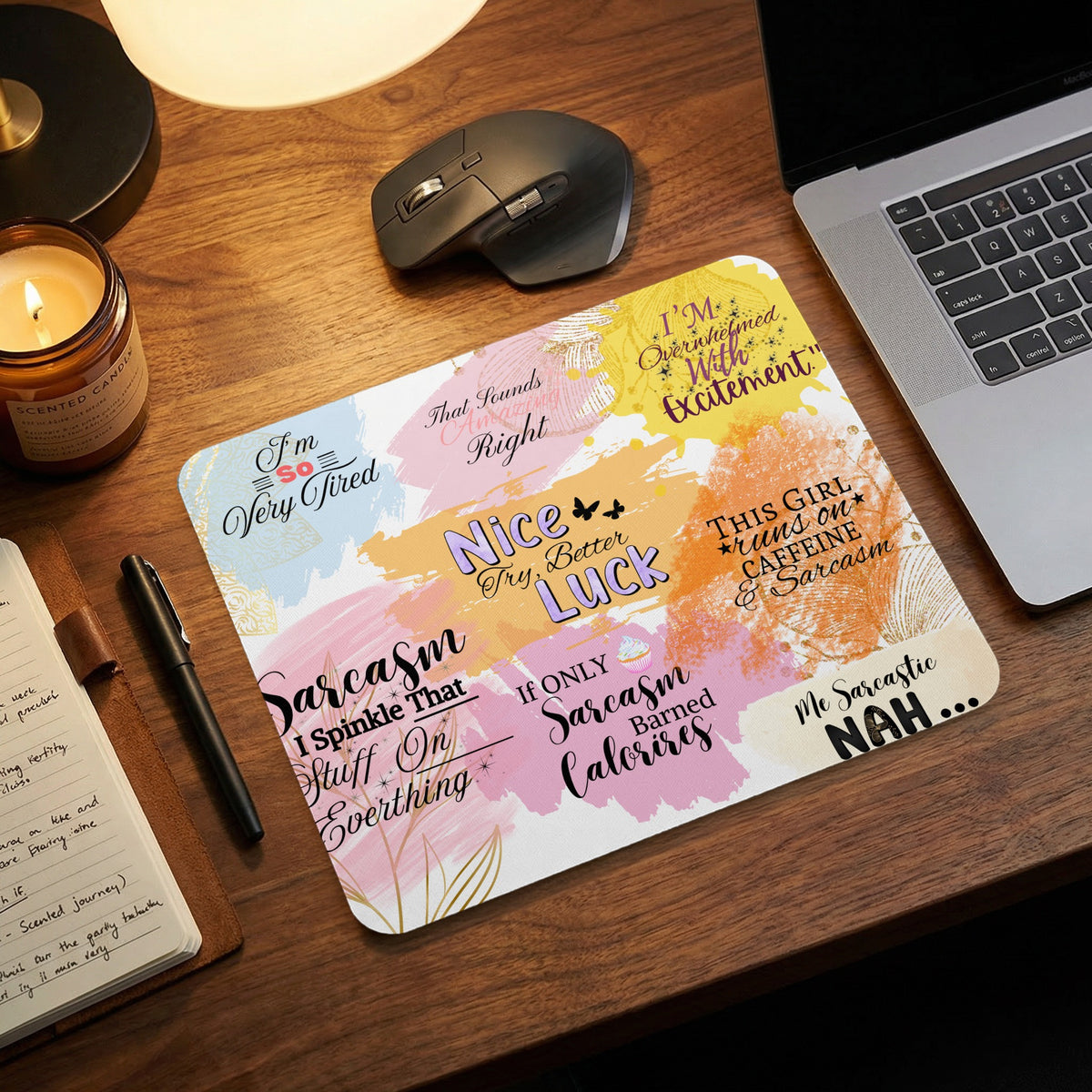 A colorful sarcastic mouse pad rests on a wooden desk next to a laptop and candle.