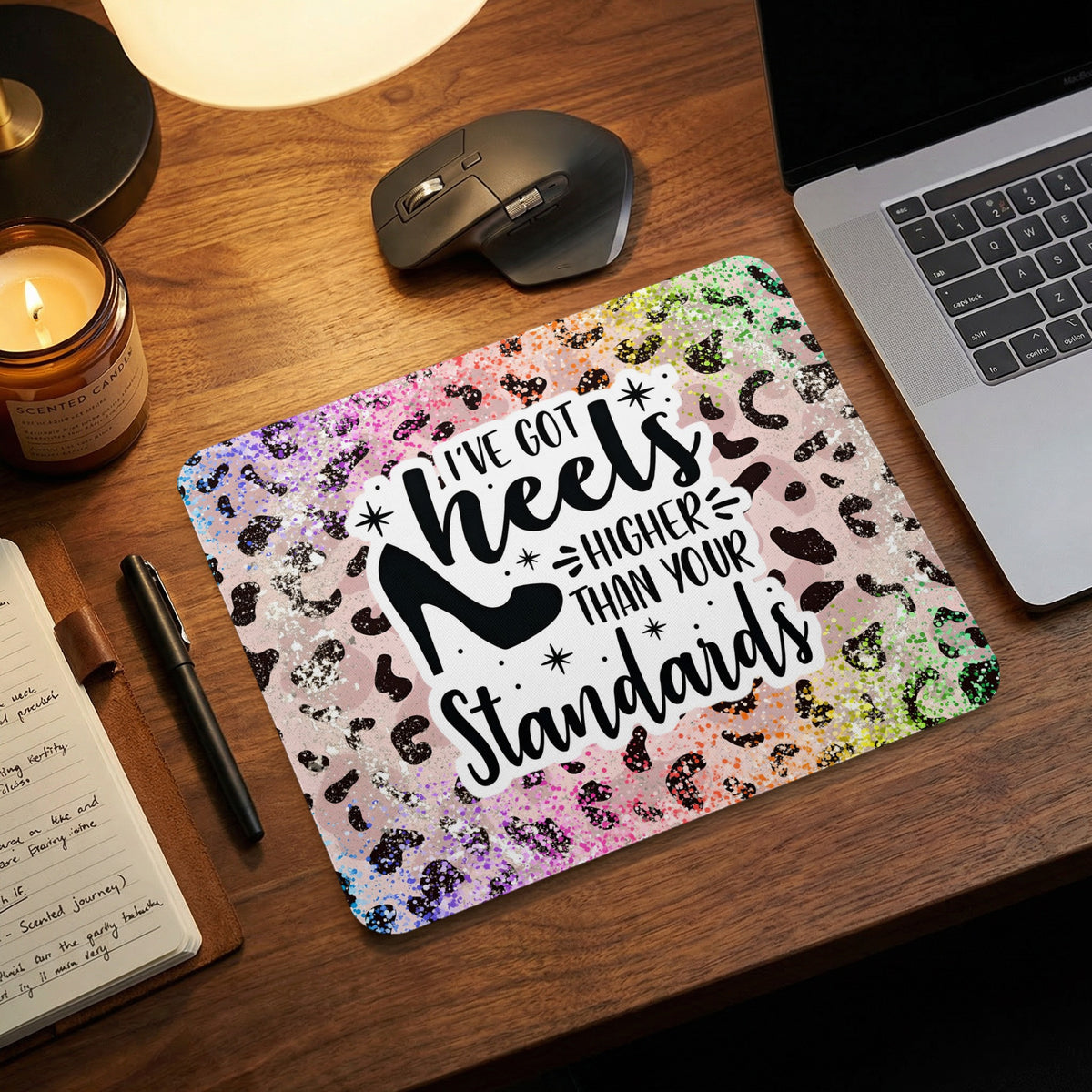 A colorful, leopard-print mouse pad with the text I've Got Heels Higher Than Your Standards lies on a wooden desk next to a laptop, mouse, and notebook.