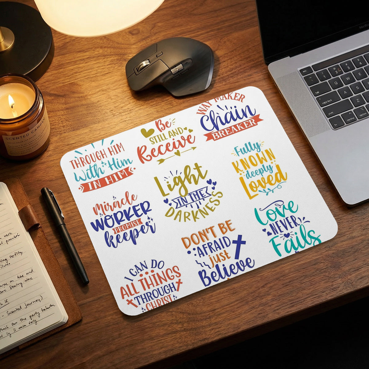 A white mousepad featuring colorful religious phrases sits on a wooden desk next to a laptop, mouse, and notebook.