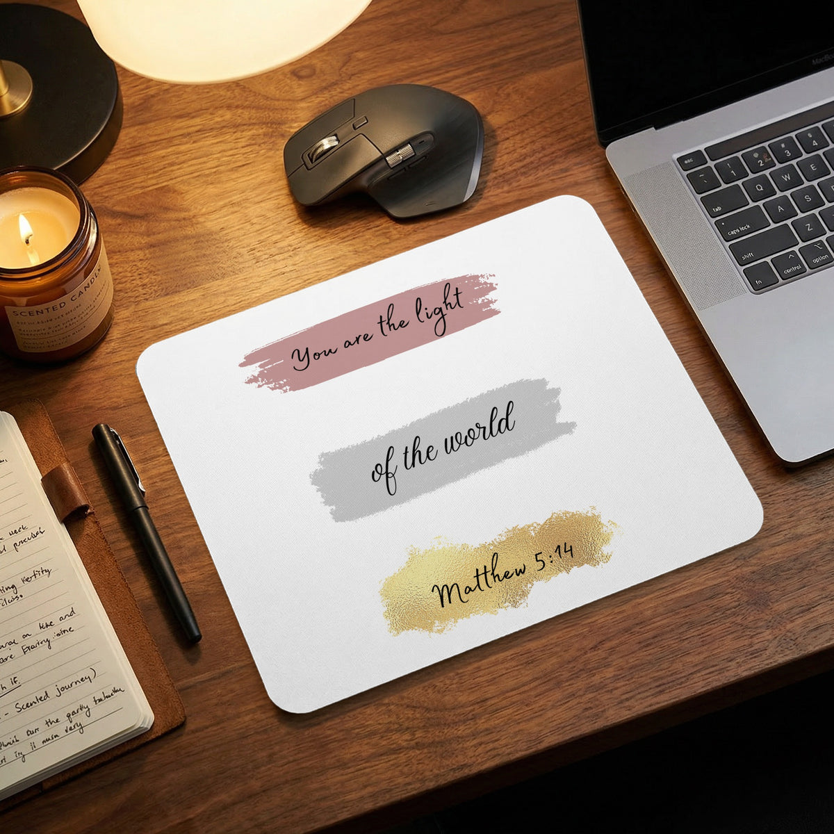 A white mousepad featuring the text You are the light of the world, Matthew 5:14 rests on a wooden desk next to a laptop, mouse, and candle.