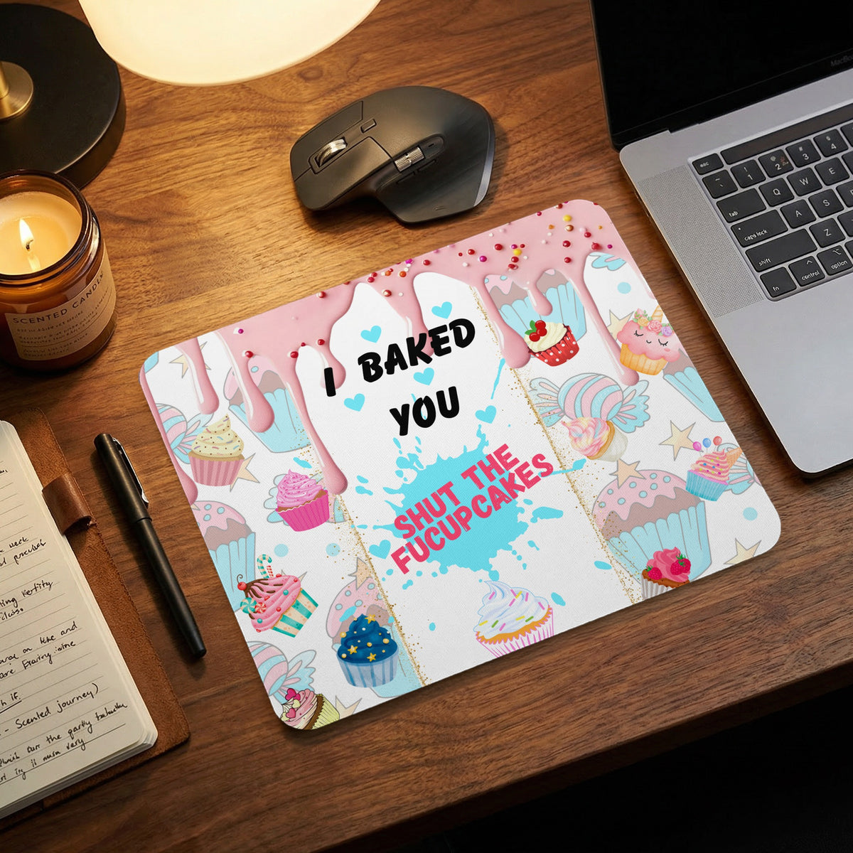 A rectangular mousepad featuring a cupcake and sprinkles pattern displays the text I BAKED YOU SHUT THE FUCUPCAKES on a wooden desk with a laptop and mouse.