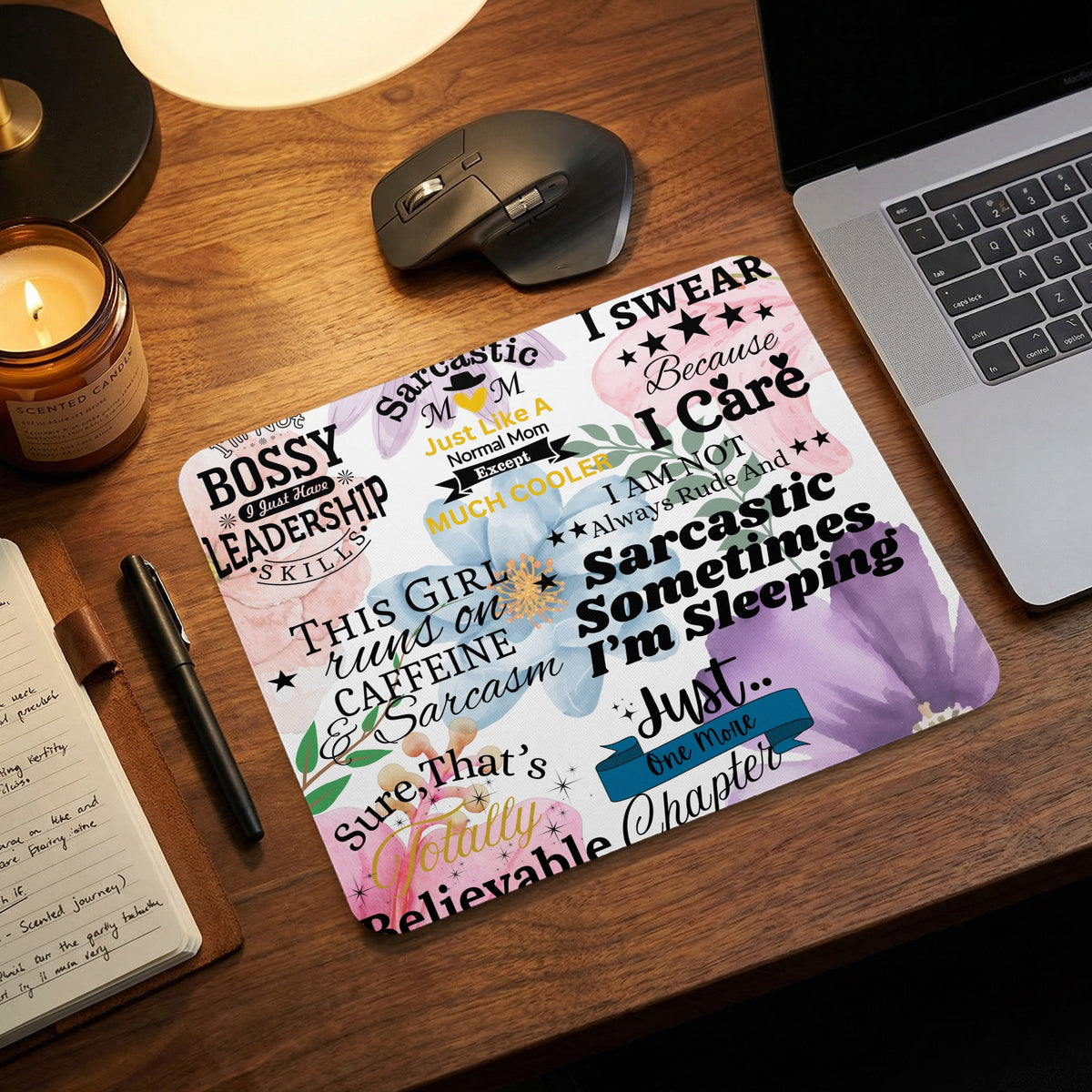 A floral mousepad featuring humorous quotes sits on a wooden desk next to a laptop, mouse, and candle.