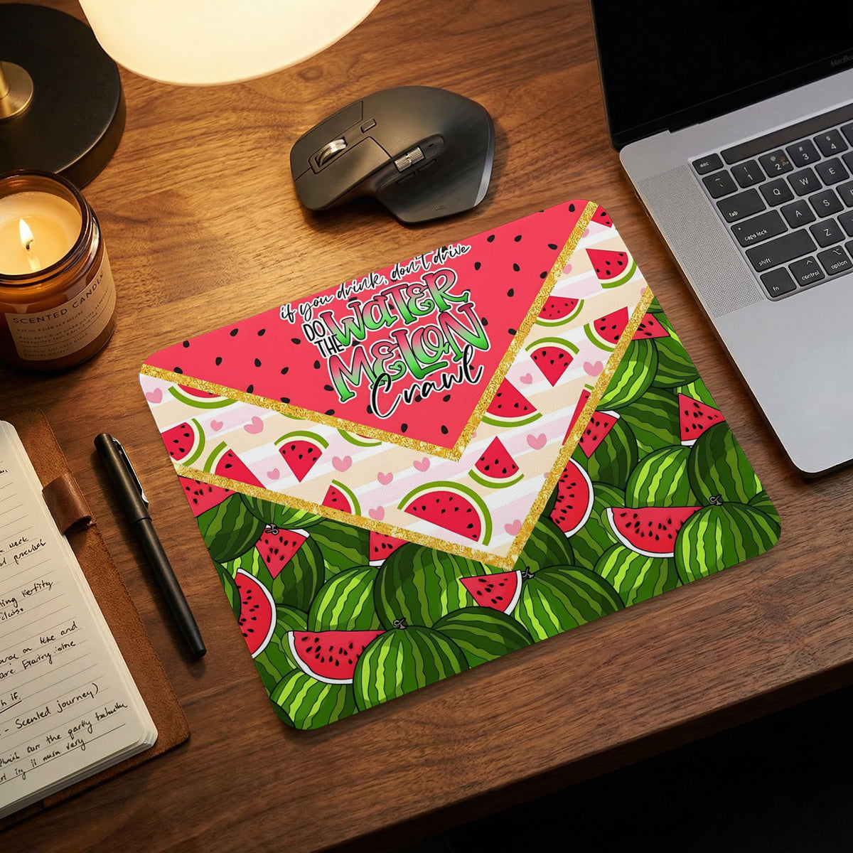 A rectangular mouse pad with a vibrant watermelon theme rests on a wooden desk near a laptop.