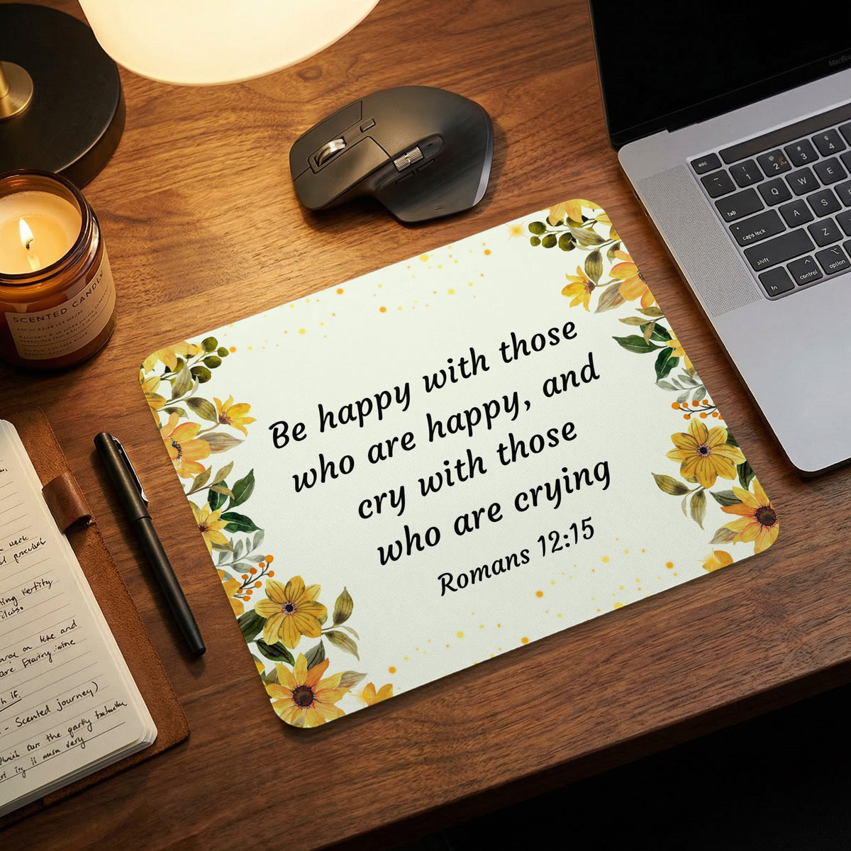 A rectangular mousepad featuring yellow floral borders and the biblical quote Romans 12:15 on a wooden desk.