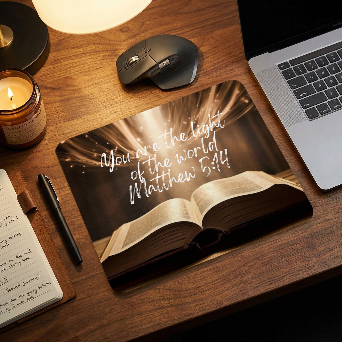 An open Bible mouse pad featuring the text You are the light of the world Matthew 5 14 rests on a wooden desk.
