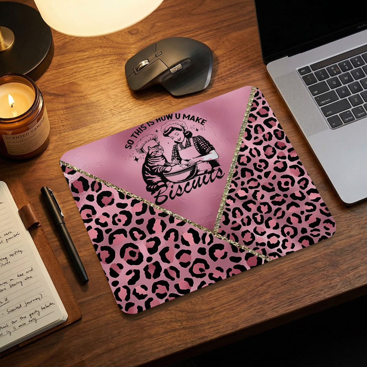 A pink mousepad with a cat baking design and leopard print borders, displayed on a wooden desk.