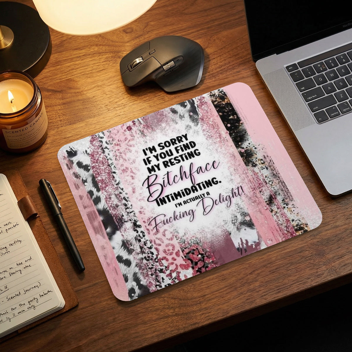 Pink and black patterned mouse pad with humorous text on a wooden desk next to a laptop.