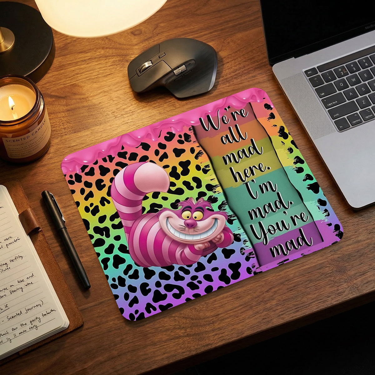 A deskmat featuring the Cheshire Cat and the quote We re all mad here on a colorful background.