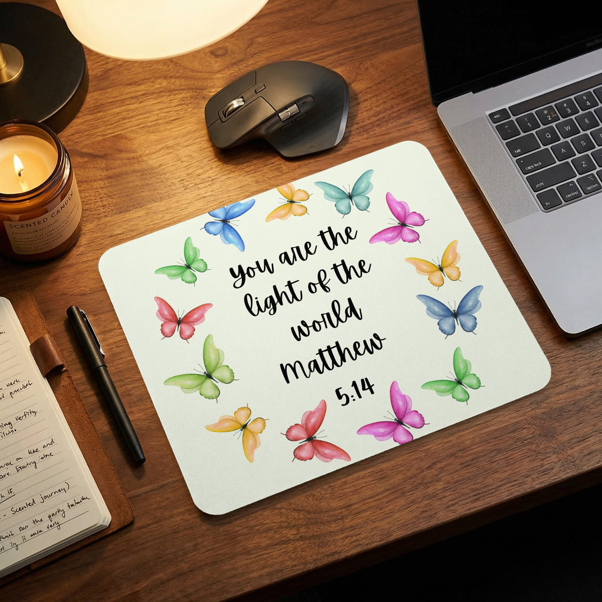 A white mousepad decorated with colorful watercolor butterflies and the text You are the light of the world Matthew 5:14 rests on a wooden desk.