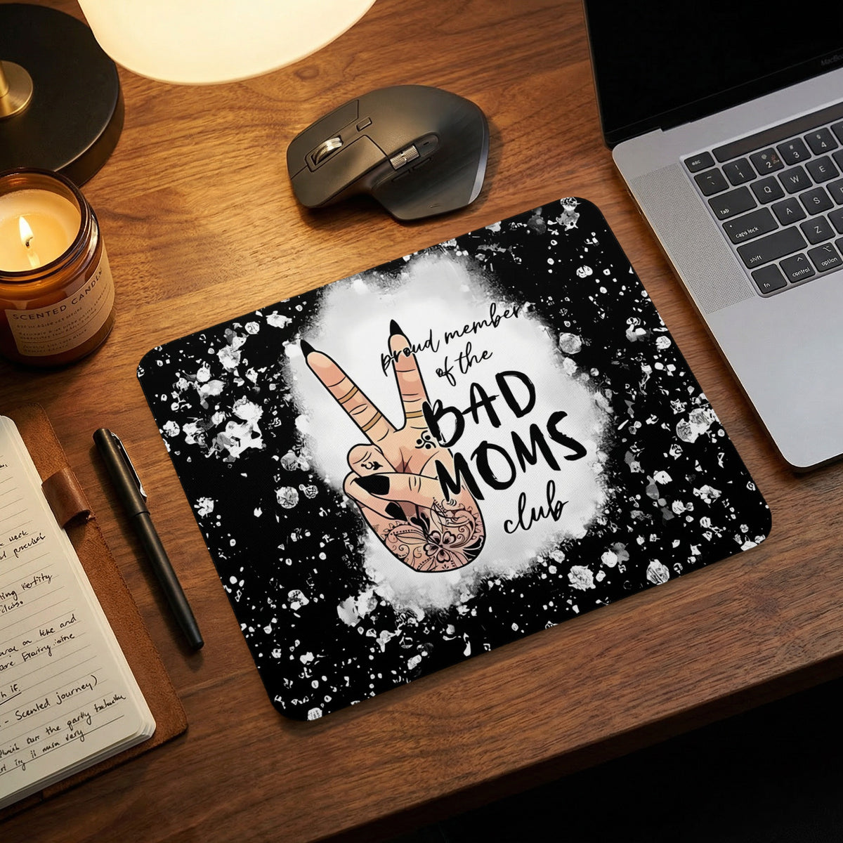 A black and white mouse pad with a hand making a peace sign and text reading proud member of the BAD MOMS club on a wooden desk.