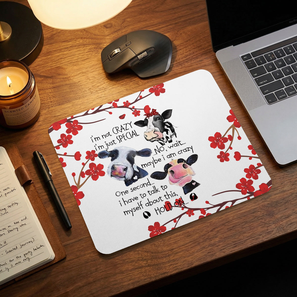 A rectangular mousepad featuring illustrations of three cows and humorous text surrounded by red cherry blossoms on a wooden desk next to a laptop.