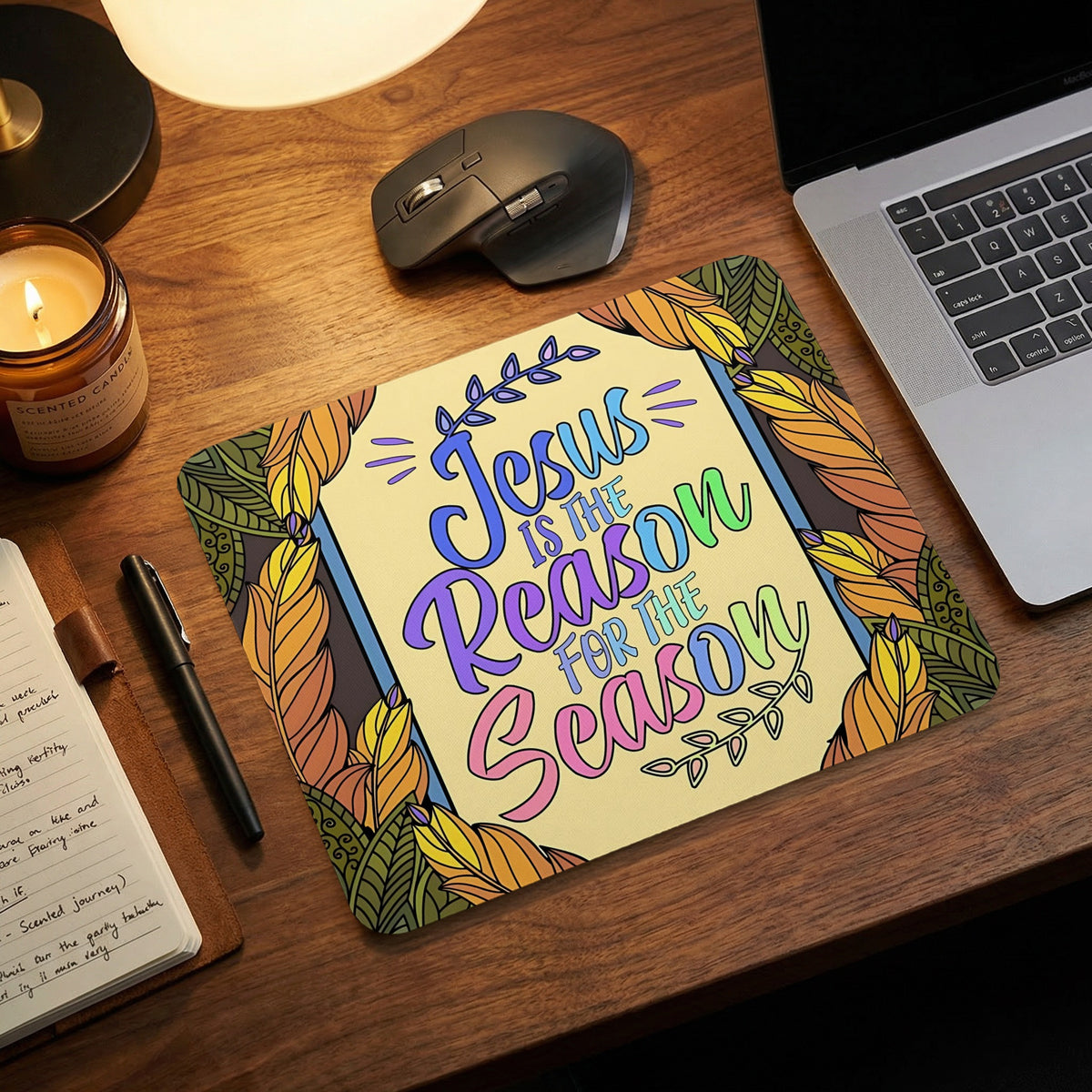 A rectangular mousepad featuring colorful leaf illustrations and the text Jesus Is the Reason for the Season sits on a wooden desk with a laptop, mouse, candle, and notebook.