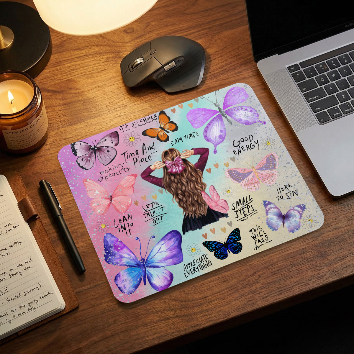 A mousepad featuring butterflies and an illustration of a woman is placed on a wooden desk near a laptop, mouse, and candle.