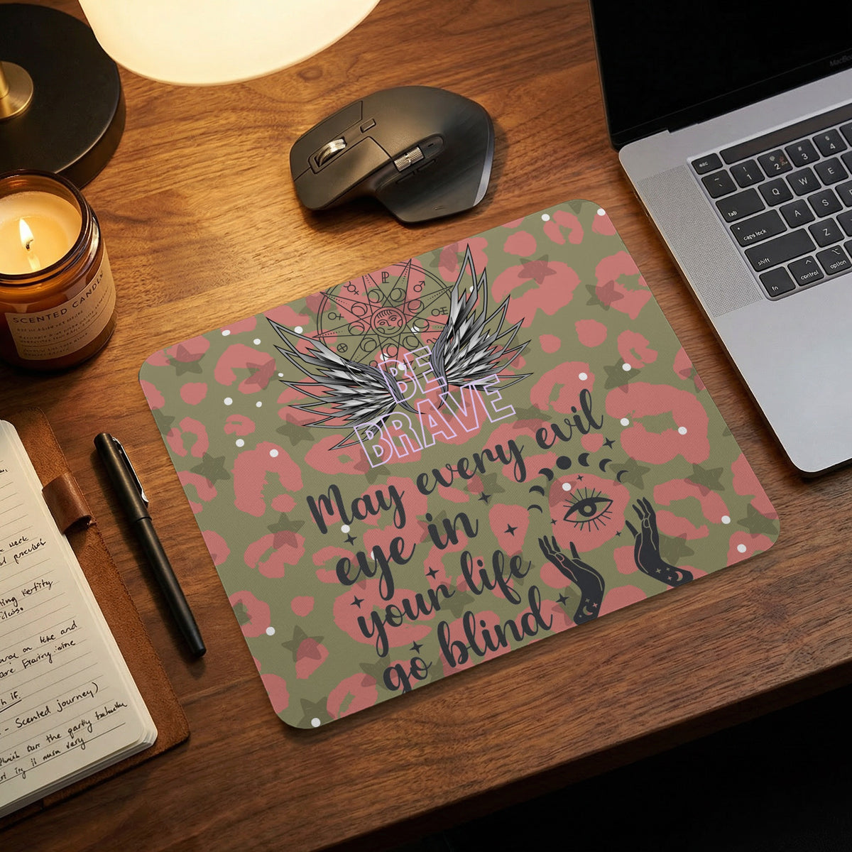 A rectangular mousepad with a pink and green leopard print features wings, mystical symbols, and the text "BE BRAVE. May every eye in your life go blind."