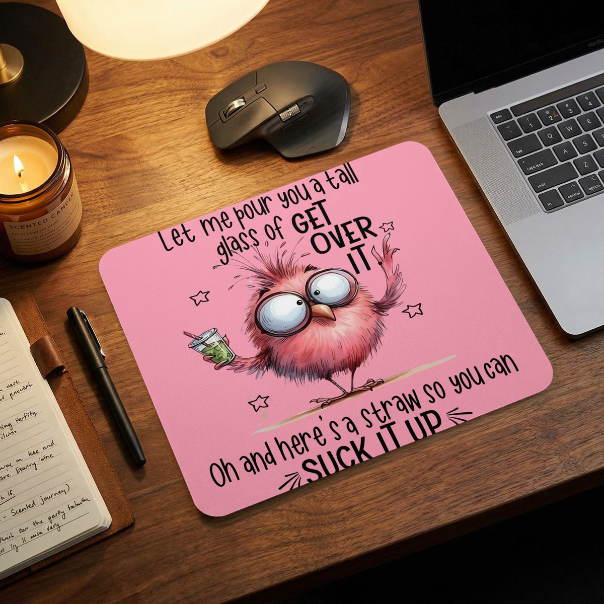 Pink mousepad with a cartoon bird, text, and desk items including a laptop, mouse, and candle.