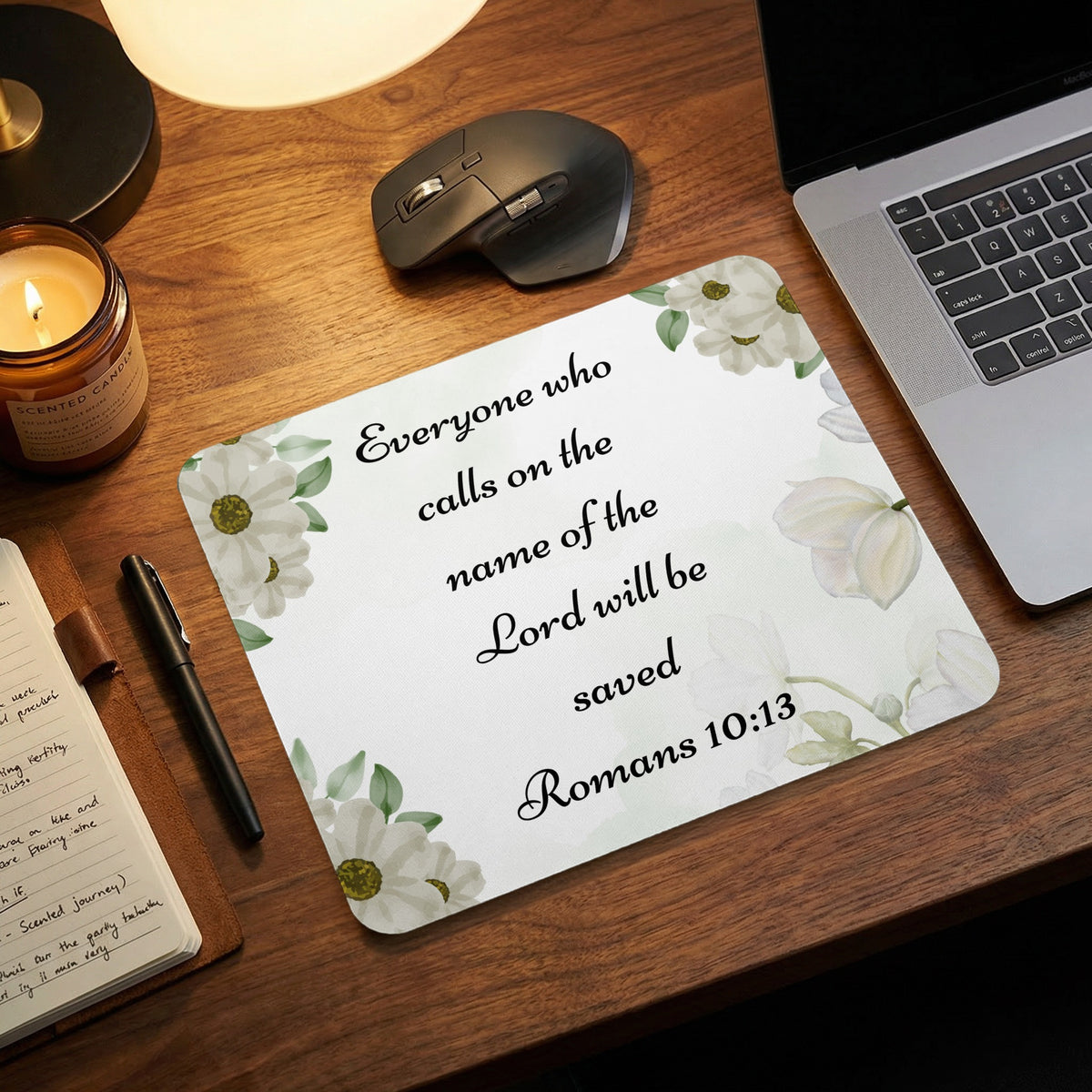 White mousepad with floral corners and the black text Everyone who calls on the name of the Lord will be saved Romans 10:13 on a wooden desk next to a laptop.