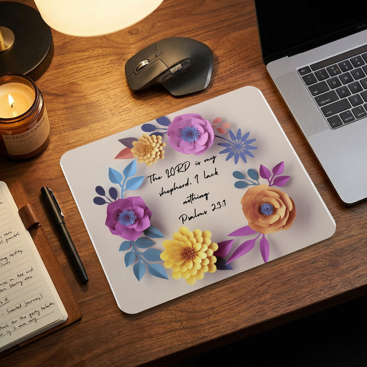 A light-colored rectangular mousepad with a floral wreath design and the text The Lord is my shepherd. I lack nothing. Psalms 23:1 rests on a wooden desk.