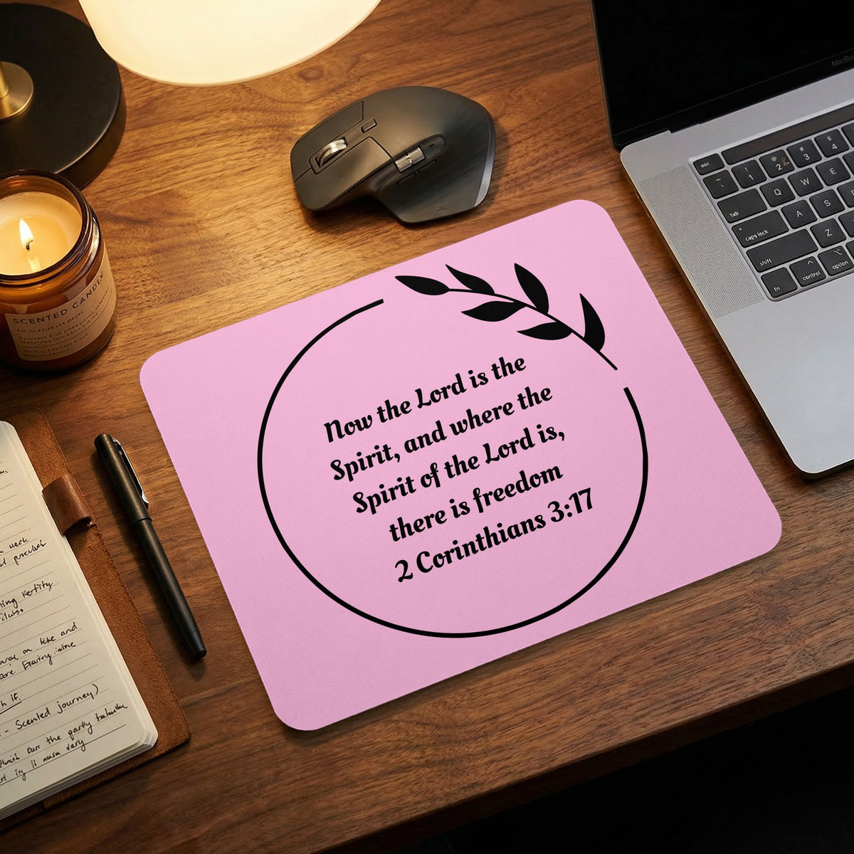 Pink rectangular mouse pad on a wooden desk featuring the Bible verse 2 Corinthians 3:17 inside a black circle.