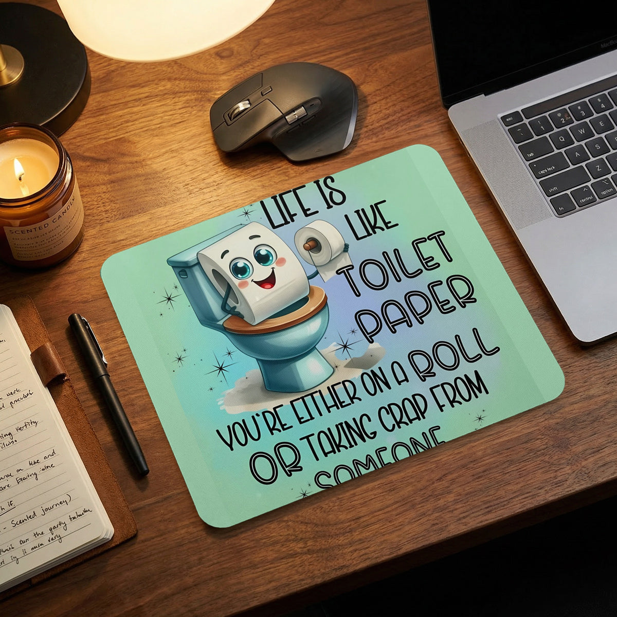 A mint green mousepad on a wooden desk features a cartoon toilet paper roll holding a smaller roll, accompanied by humorous text.