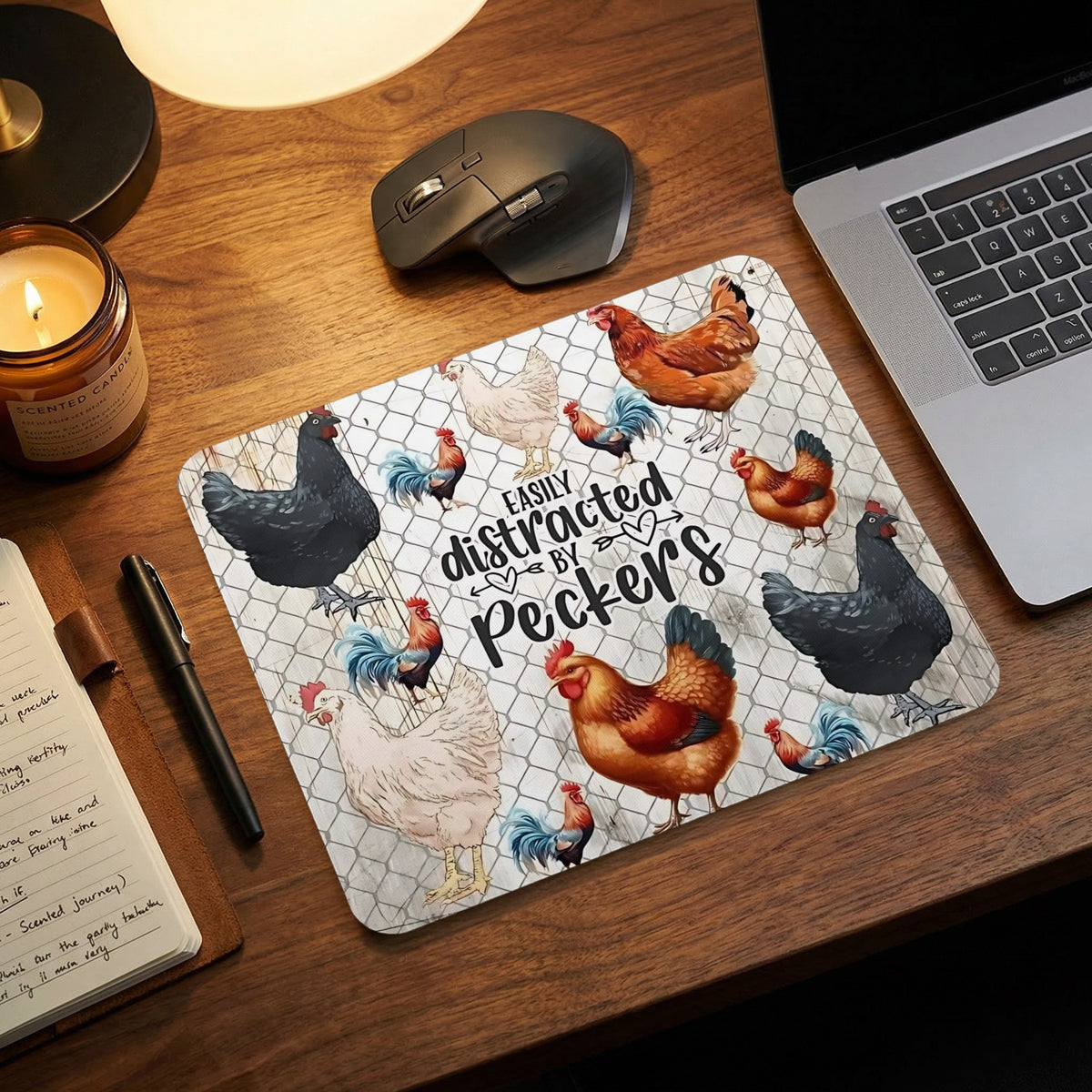 Chicken-themed mousepad on a wooden desk next to a laptop, mouse, candle, and notebook.