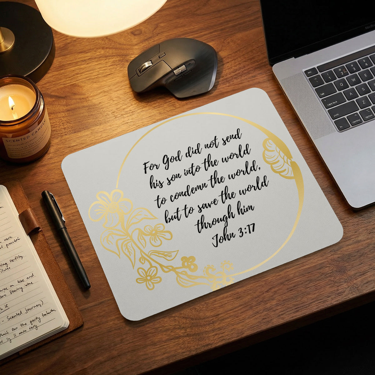 A light gray mousepad featuring a gold floral wreath and John 3:17 Bible verse on a wooden desk.