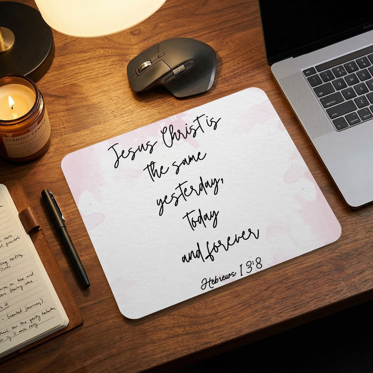 A rectangular mousepad with pink watercolor accents displaying the text Jesus Christ is the same yesterday, today and forever, Hebrews 13:8 sits on a wooden desk next to a laptop, mouse, notepad, and candle.