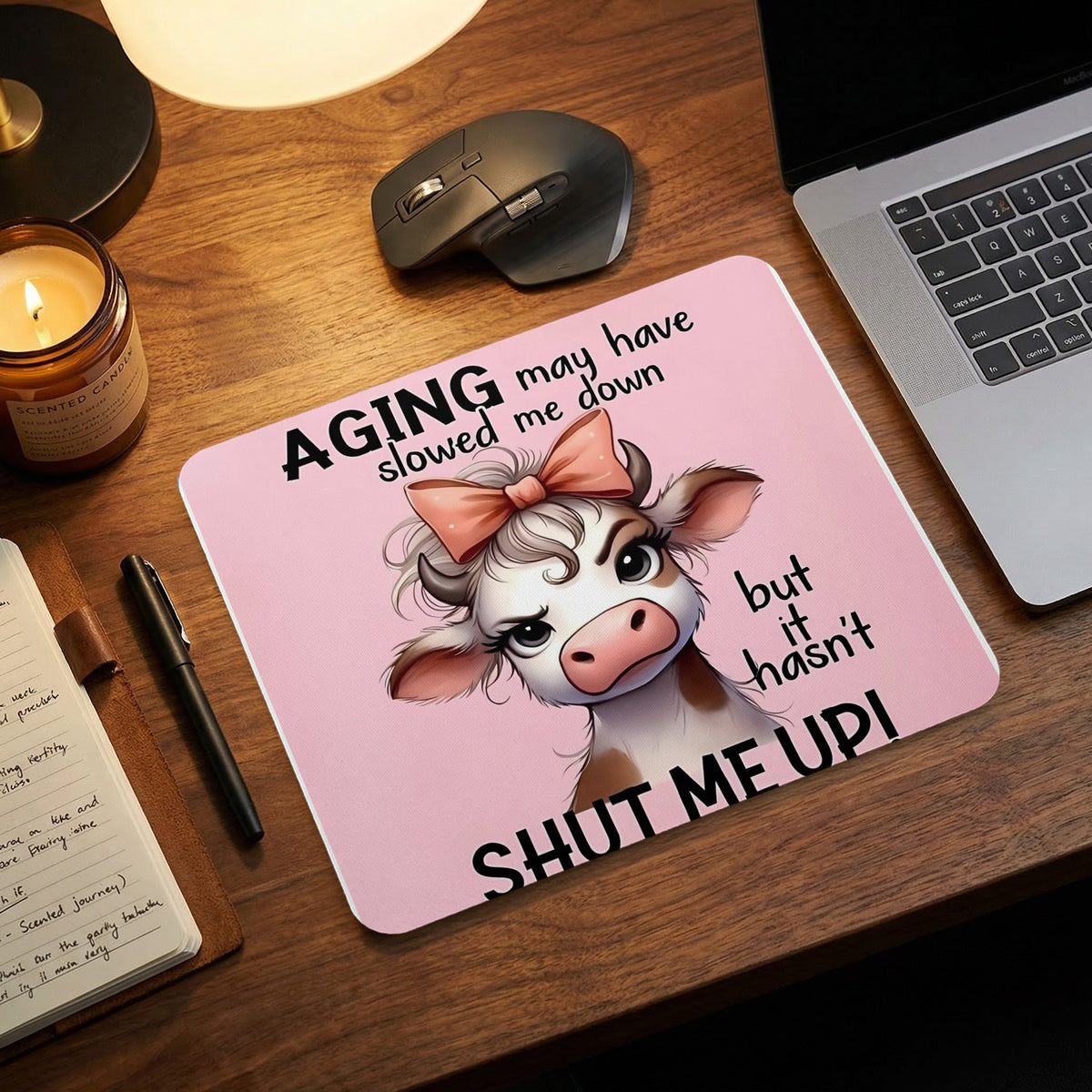 A pink mouse pad featuring an illustrated cow and the text AGING MAY HAVE SLOWED ME DOWN BUT IT HASNT SHUT ME UP on a wooden desk.